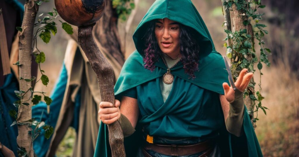 A woman in a green cloak and hood, holding a stick, embodies the essence of a Druid in a roleplay setting.