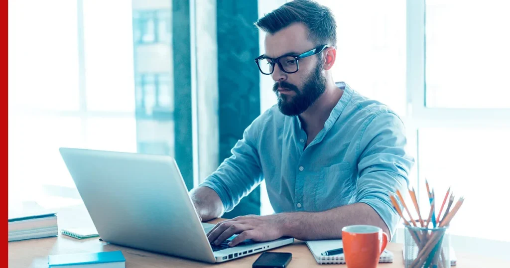 A man with glasses and a beard is focused on his laptop, embodying the essence of letting your personal style prevail.
