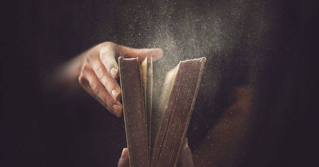 A person holds a dusty book titled "Books of Prophets," with dust particles swirling in the air around it.