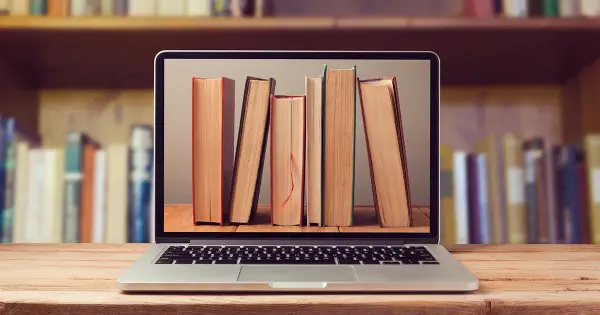 A laptop with books on it rests on a table, positioned in front of a bookshelf, illustrating "Ebooks Don’t Work That Well for Some Categories.